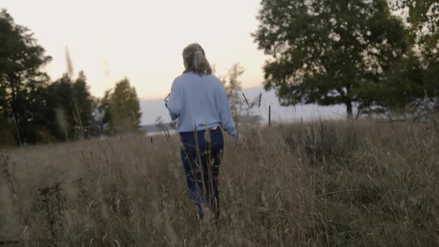 A woman walks through a clearing of tall grass with trees in the distance on both sides of her