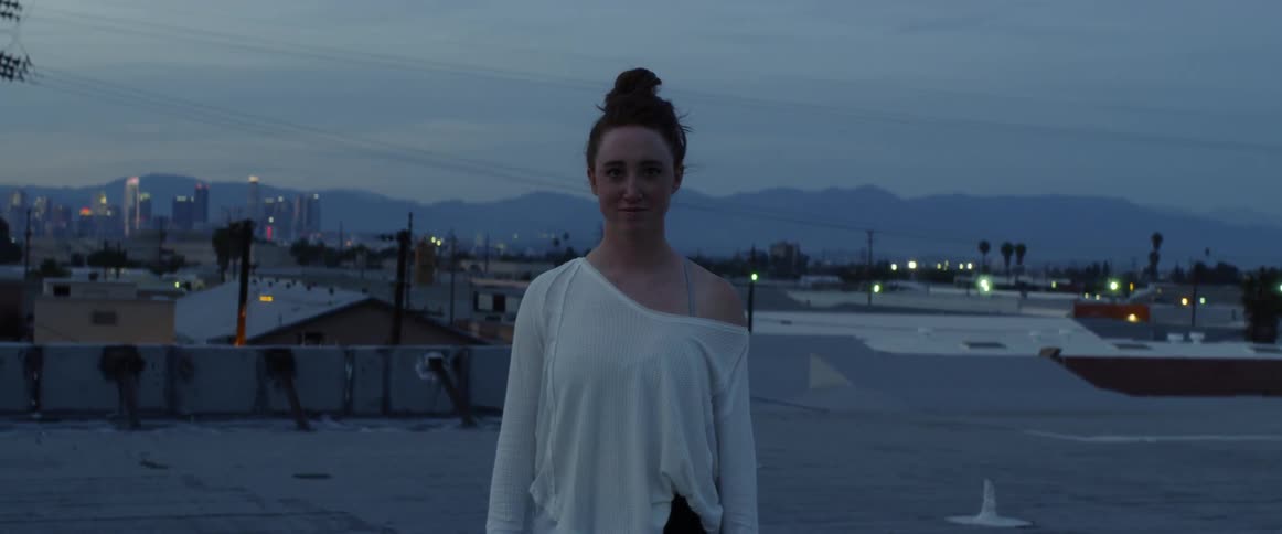 A woman in a white shirt stands smiling at us with the city behind her