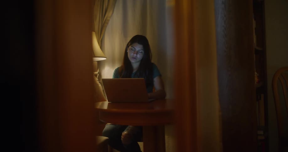 A young lady sits typing on a laptop at a table in a room lit by a table lamp