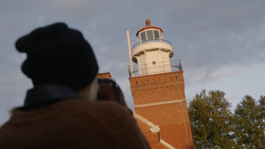 A woman takes a photo of a lighthouse then checks the shot and smiles