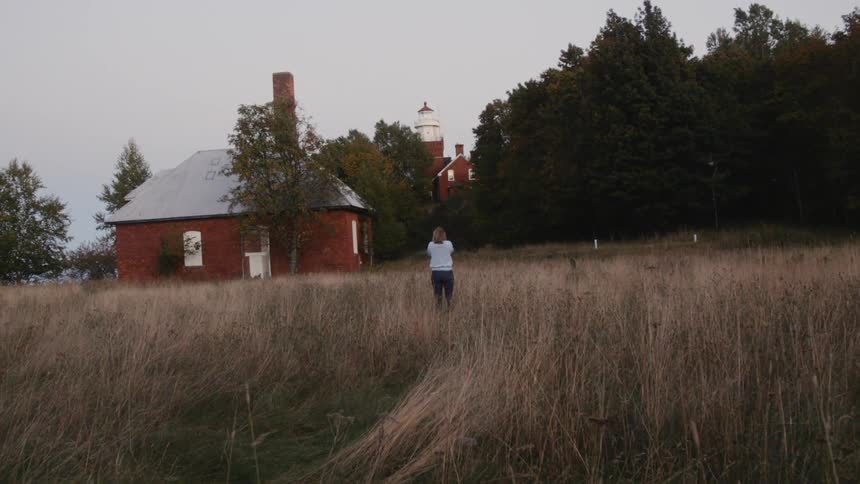 A woman stands in a field near an old red building at dusk