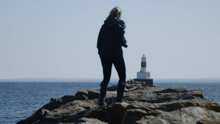 A woman walks along a rocky path that leads to a lighthouse