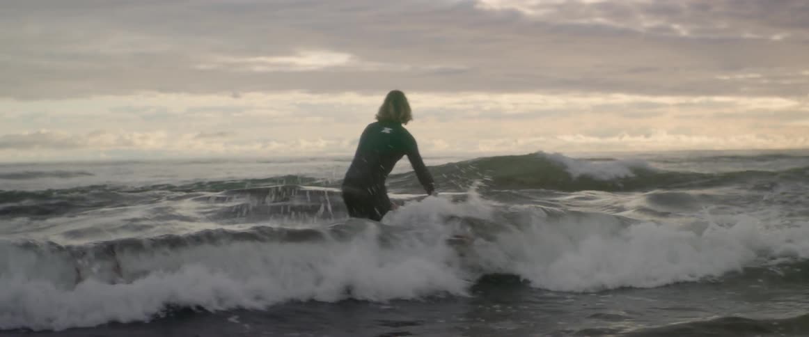 A person in a wet suit walks a surfboard into the ocean and holds it while waves go under it then lays on the surfboard and paddles deeper into the ocean