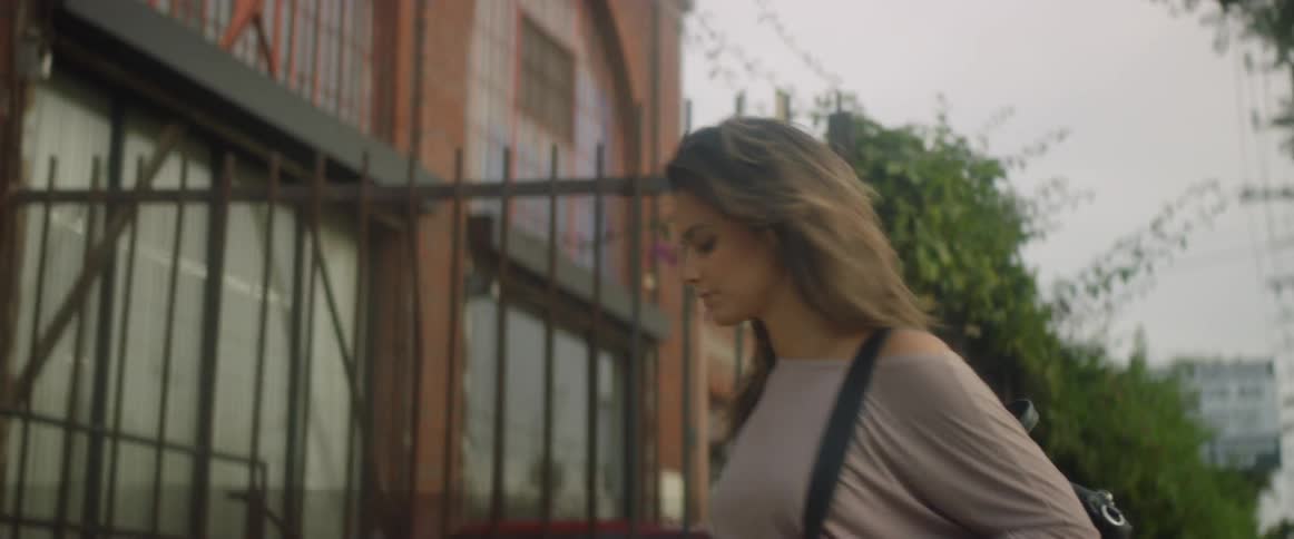 A woman wearing a backpack is walking past a fence toward a building entrance in daylight