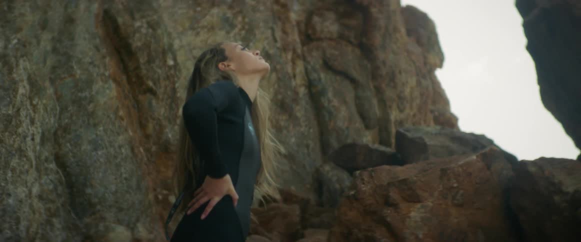 A woman in a wet suit stands on a cliff and looks into the distance