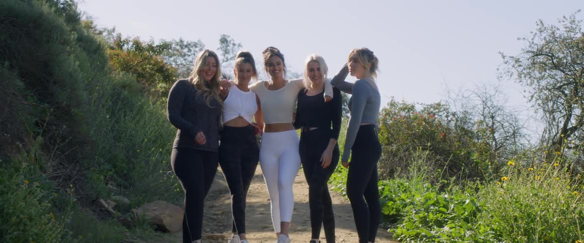 Five woman stand in a line on a path smiling while they hold onto each other