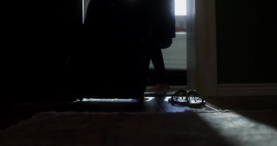A person wheels a suitcase into a bedroom then goes to sit on the bed