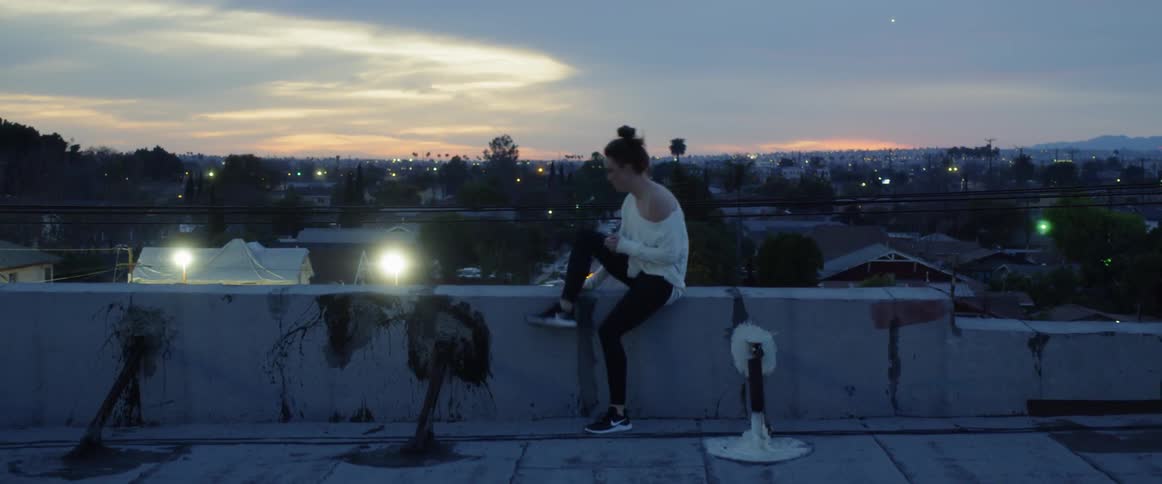 A woman walks across a rooftop to sit on a ledge and begin a phone call at dusk
