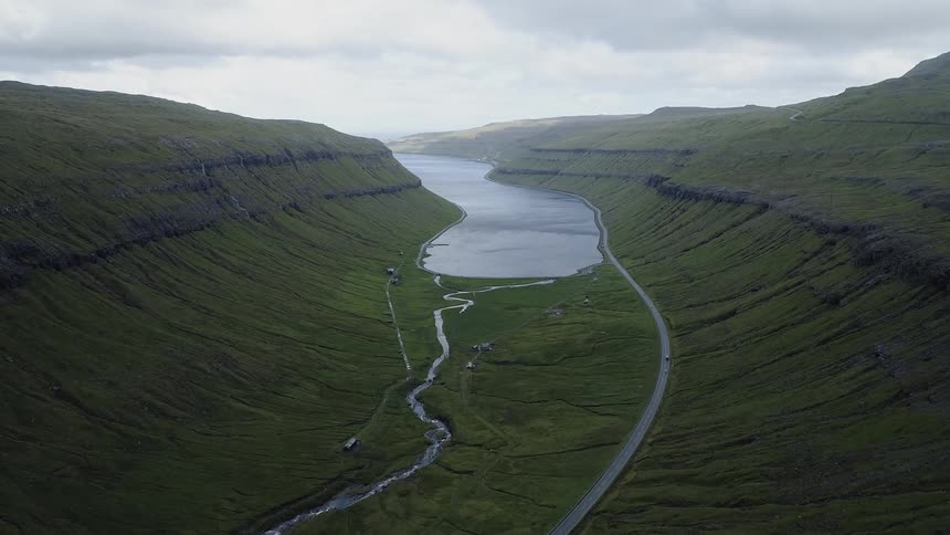 Cars driving down a road alongside a grassy hill next to a large river