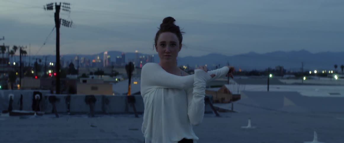 A woman in a white shirt stretches on a rooftop at dusk