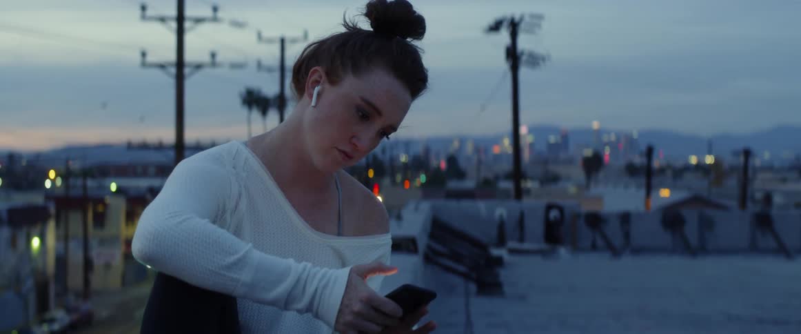 A woman with earbuds sits on a bench in the city and taps her phone before looking behind her