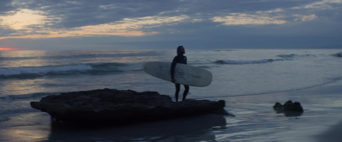 A man in a wetsuit carries his surfboard out of the ocean at sunset