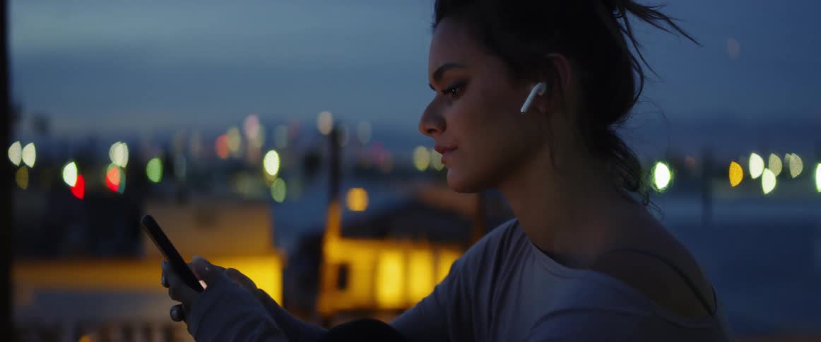 A woman scrolls on her phone at dusk as she wears wireless headphones