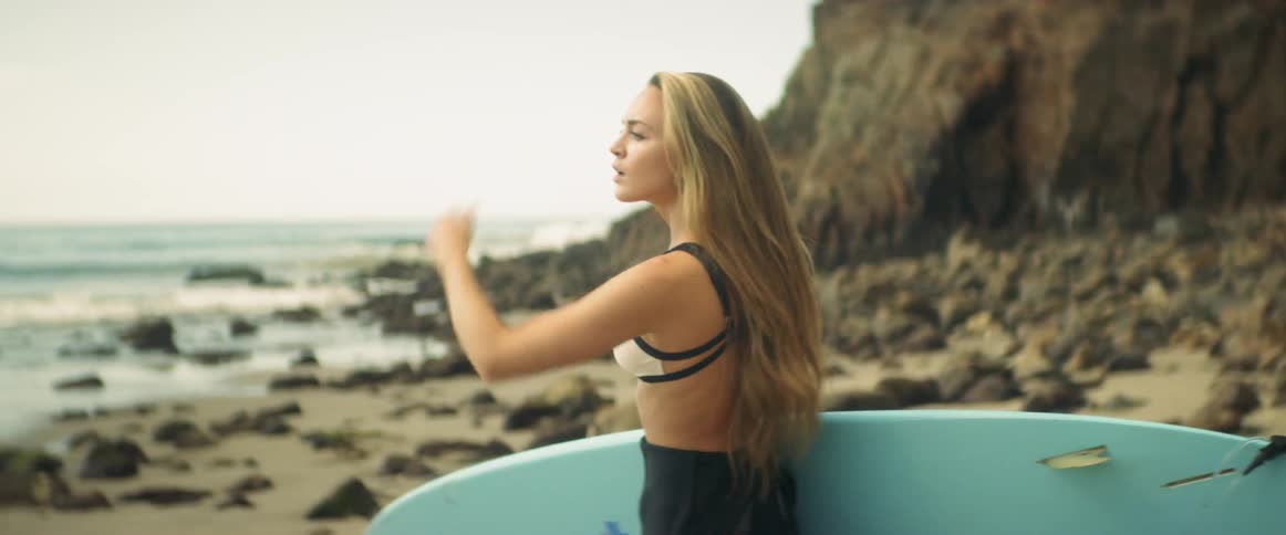 A woman holding a surfboard looks around on a rocky beach