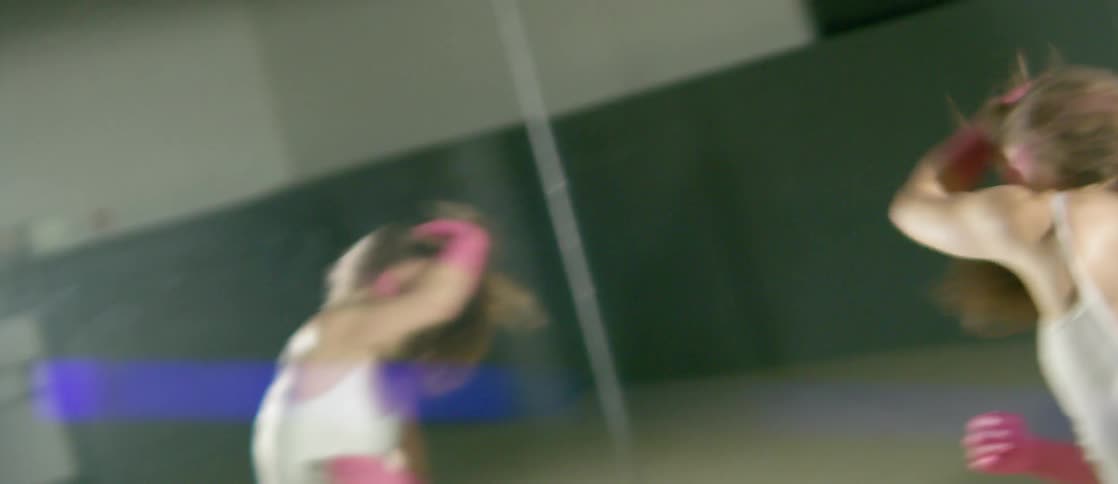 A woman dancing in front of a mirror wearing a white leotard and pink gloves