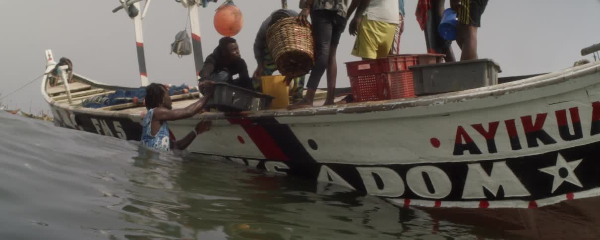 A group of men stand on a fishing boat pouring fish from a bucket while one man stands in the water