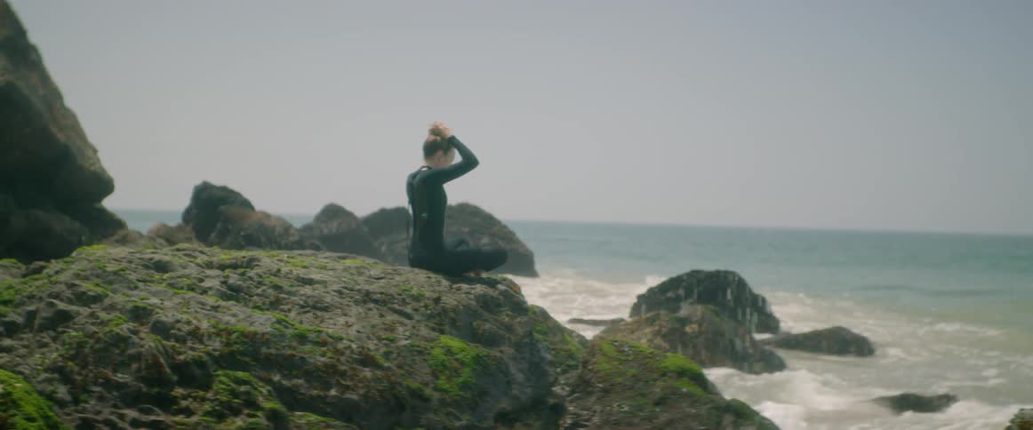 A woman in a wet suit fixes her hair while sitting on rocks by the ocean