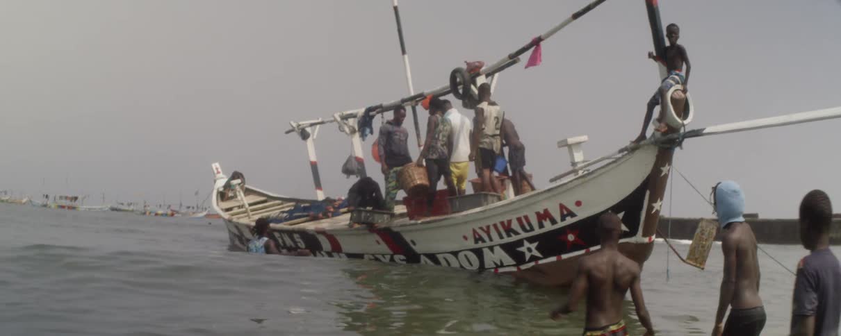 Several men stand on a boat and a few people stand in the water splashing