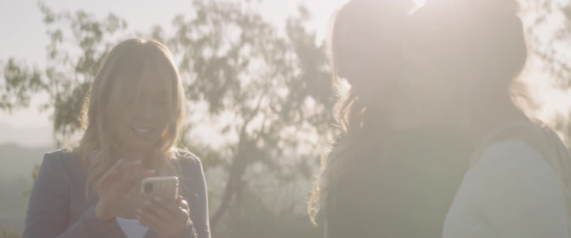 Three women stand in the sunlight with trees behind them as they laugh and look at a phone