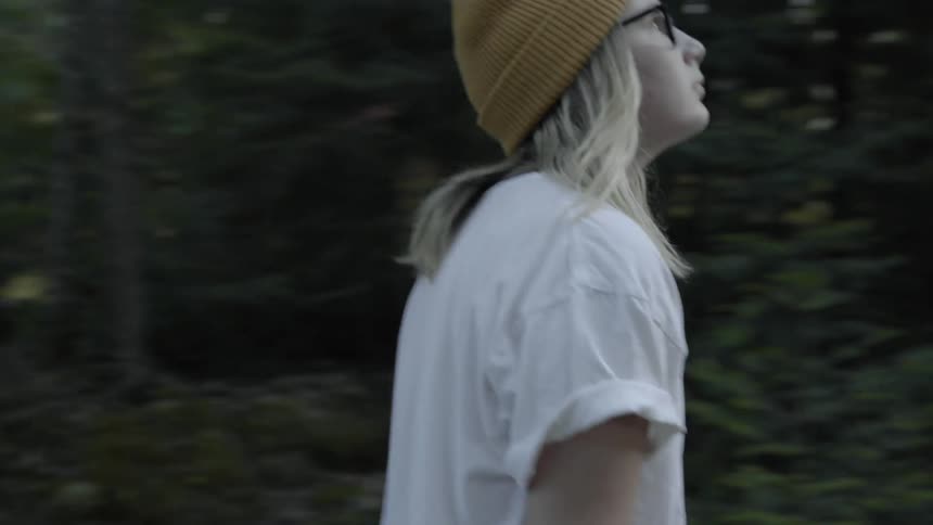 A woman wearing a beanie looking at a camera and walking through a forest