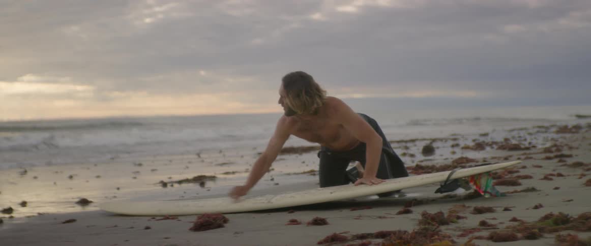 A man kneels on the sand as waves crash onto the beach while he waxes his surfboard