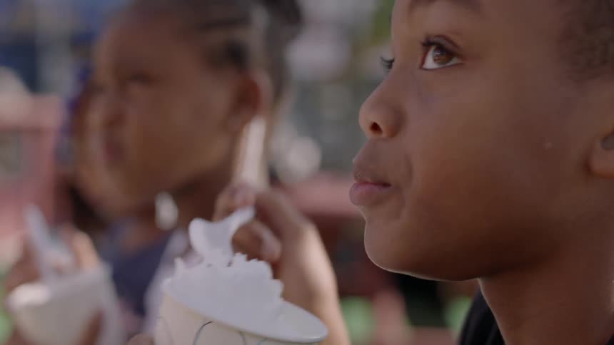 Children in a line eat a frozen desert as people sit on picnic tables behind them