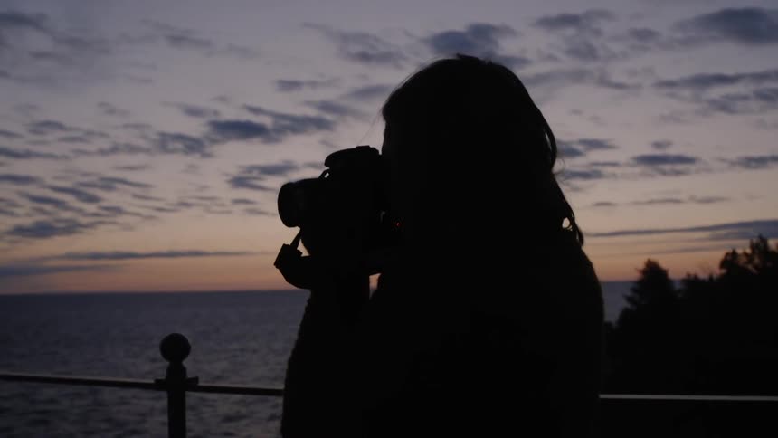 Woman taking a picture of a body of water while the sky is barely light by a barely present sun