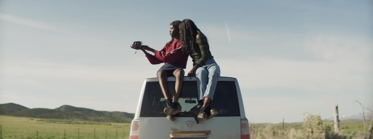 Two women sit on top of a car and record themselves with a video camera