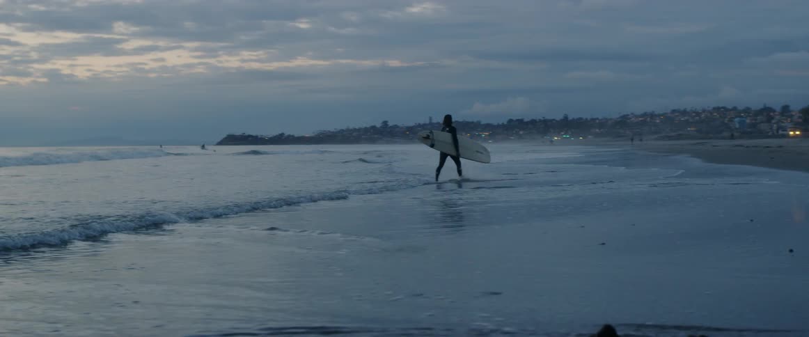 A surfer walks with their surfboard under their arm in ankle deep water