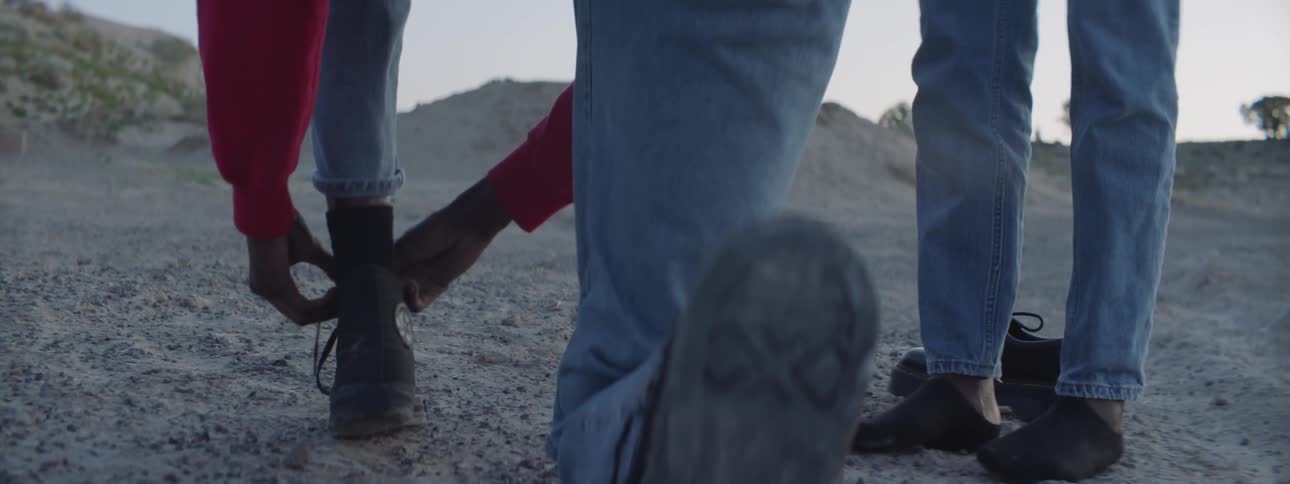 A person unties their shoe in the desert while a person wearing no shoes stands next to them