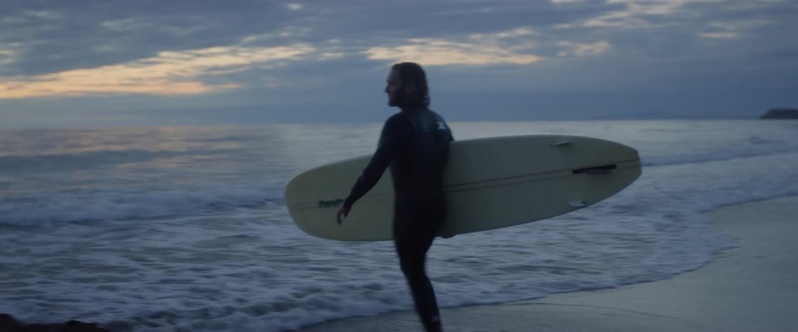 Man carrying a surfboard walks into the ocean at sunset