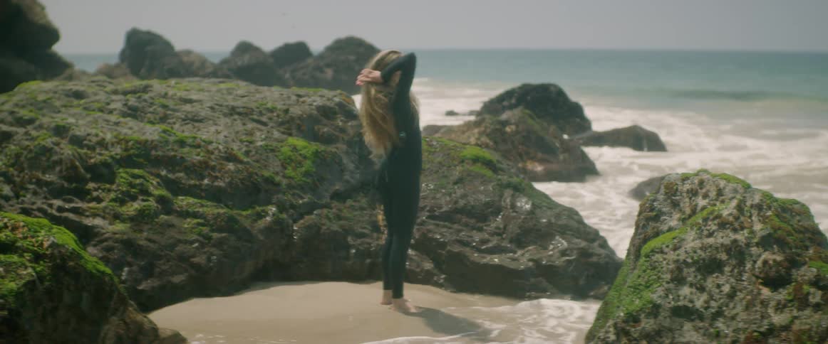 A woman in a wet suit wets her hands with water from the ocean and runs them through her hair