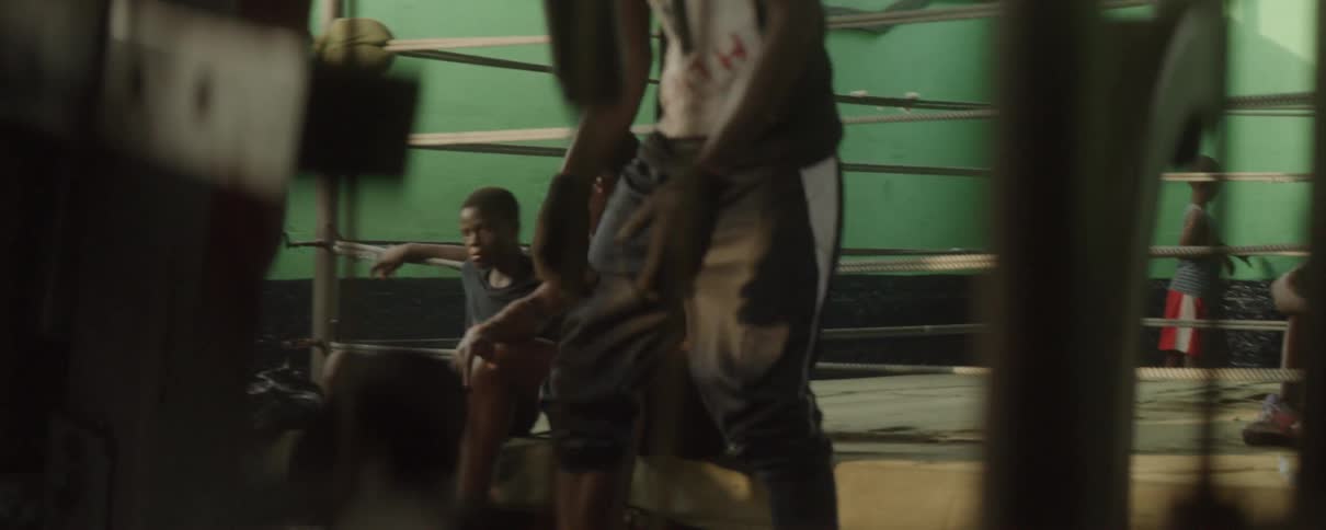 Two men sit at the edge of a boxing ring and another man flips over a large tire