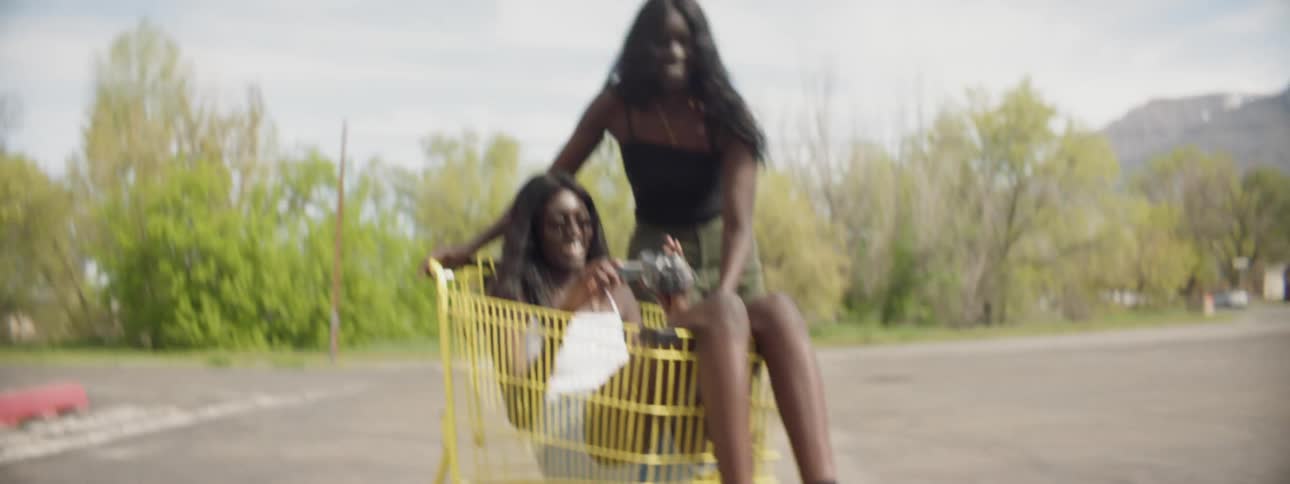A woman pushes another woman in a shopping cart through a parking lot