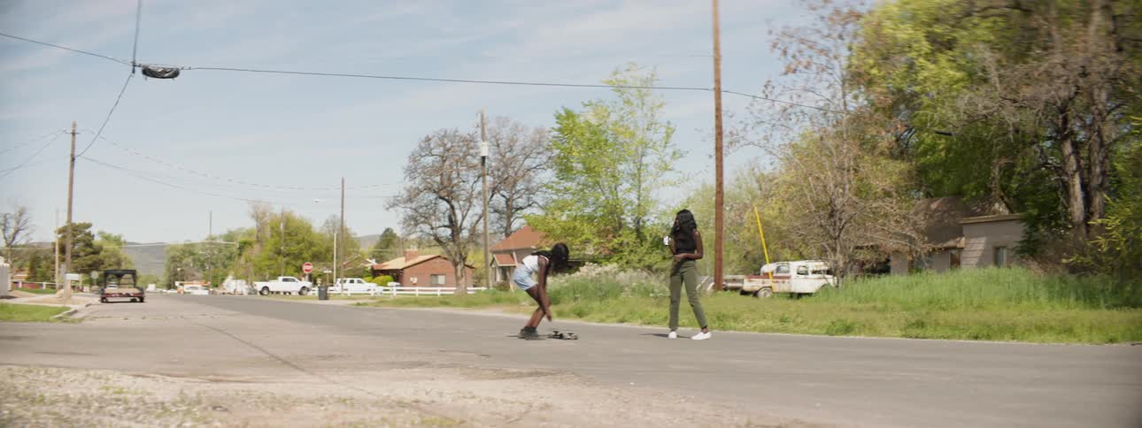 Two women stand in the middle of a rural road before one of them starts to skateboard