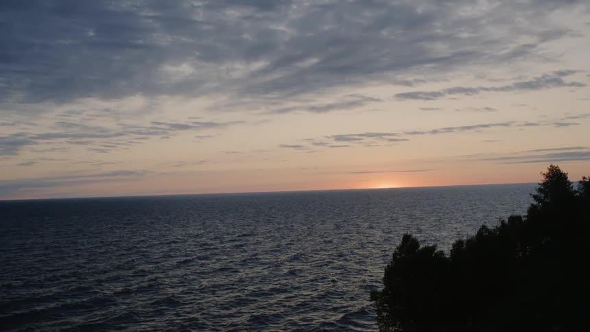 The sun sets behind the choppy ocean with pine trees to the side
