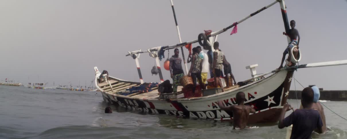 Two men walk through the water toward a boat carrying a group of men