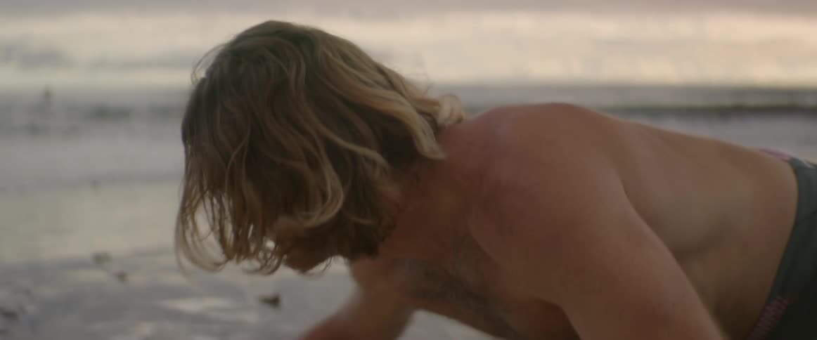 Topless man kneeling on the beach scrubbing his surfboard