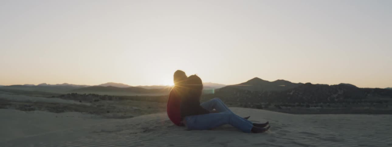 Two women sit down on the beach and then lay down together on the sand