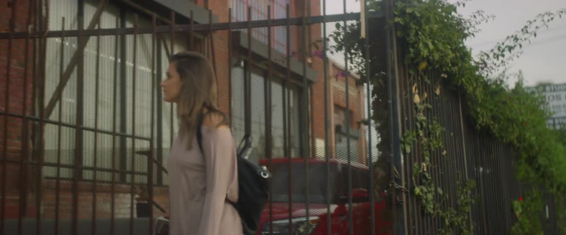 A woman walks by a metal fence with an industrial looking building behind it