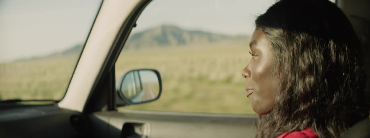 A woman rides in the passenger seat talking as the country whips by the passenger window