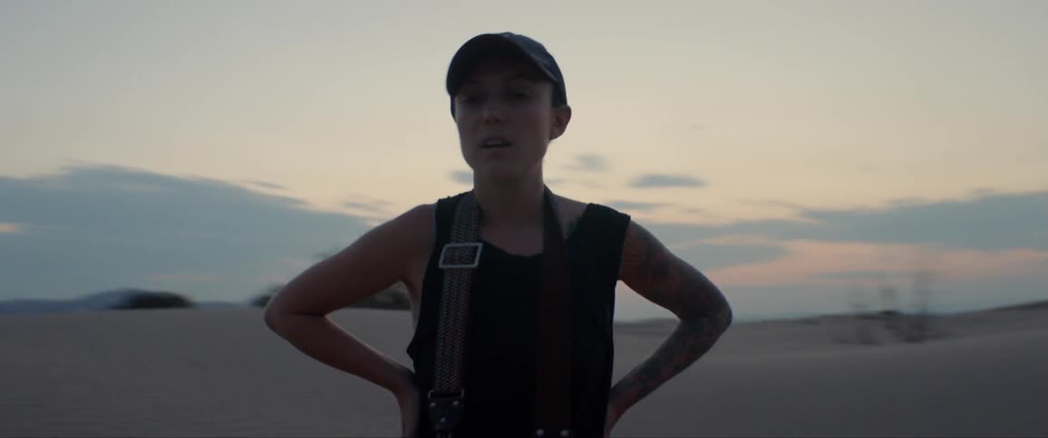 A woman wearing a hat and tank top stands in the desert with her hands on her hips