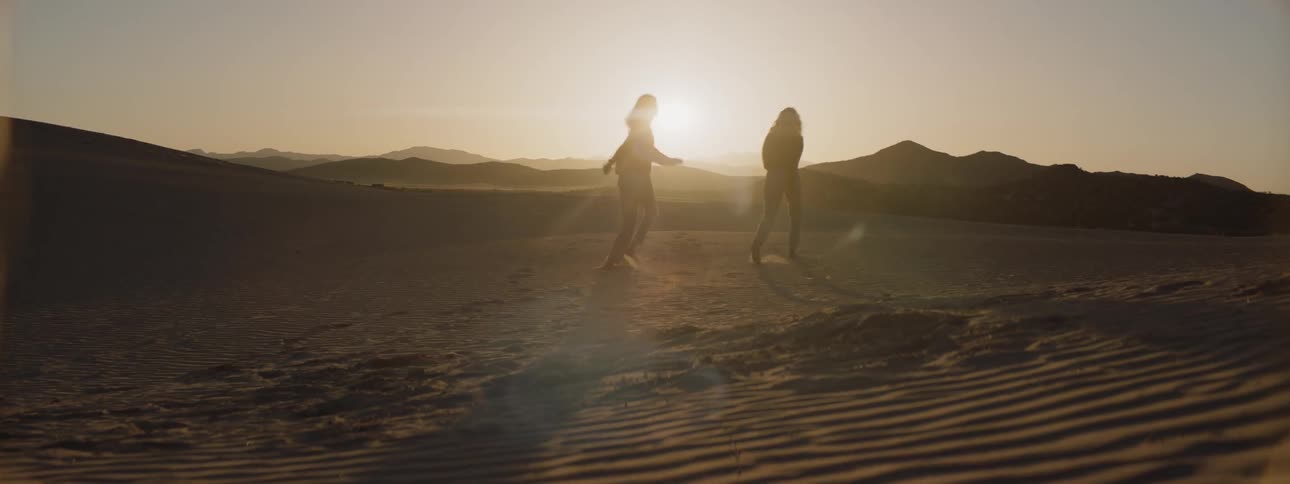 Two women skip on the sand in the setting sun