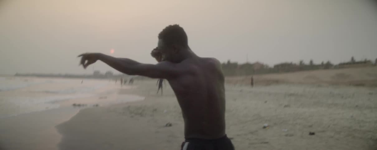 A man jumps and shadow boxes by the ocean shore as people walk behind him