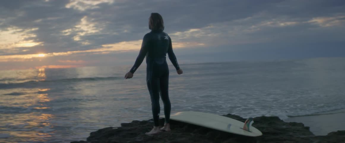 A man in a wet suit stands by his surfboard on a large rock and looks across the ocean at sunset