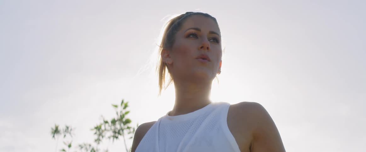 A woman in a white shirt stretches in the sun with a tree behind her