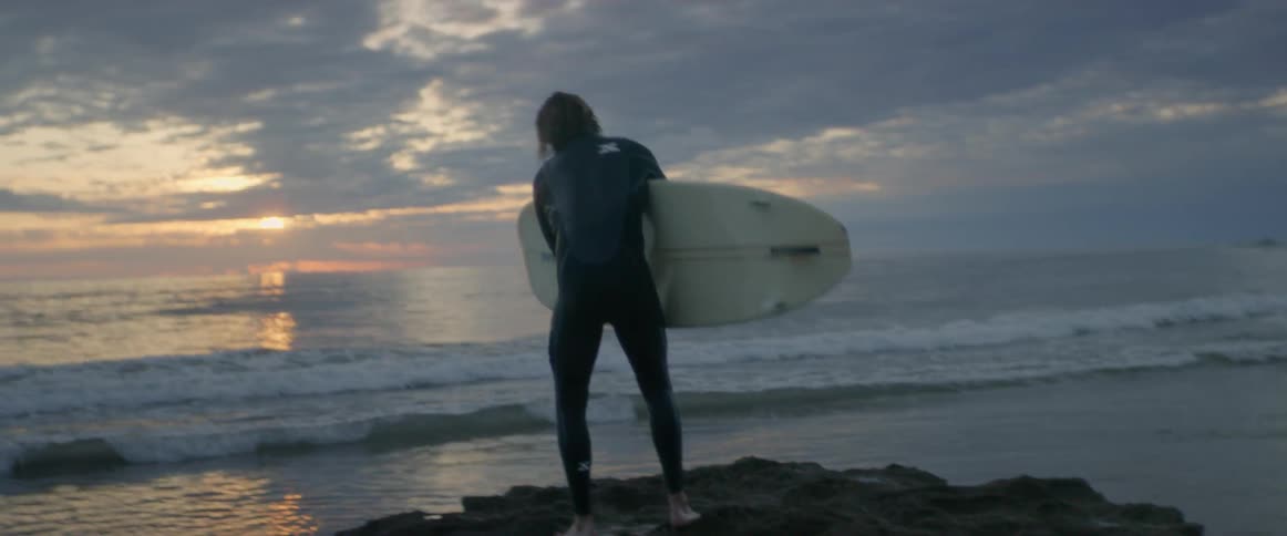 A male standing on a rock next to the sea and picking up his surfboard