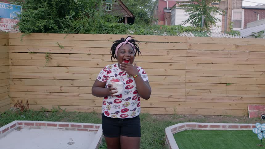A young girl eats ice cream without a spoon in a backyard and laughs