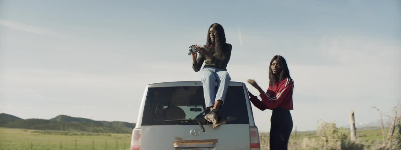 A woman sits on top of a car taking selfies as another woman paces on the ground