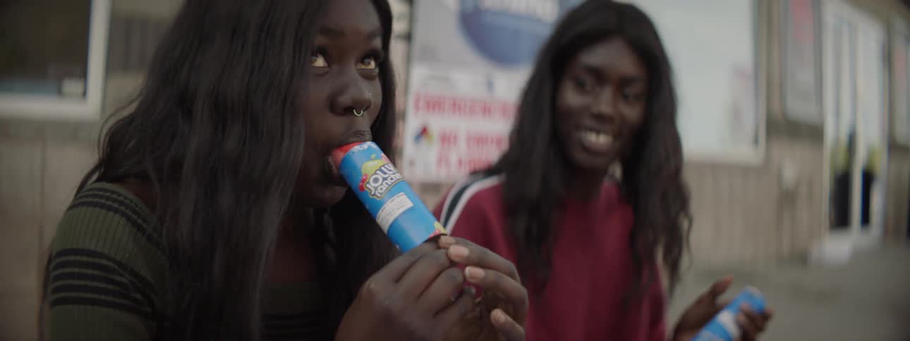 Two women eat push pops sitting on a sidewalk outside a building